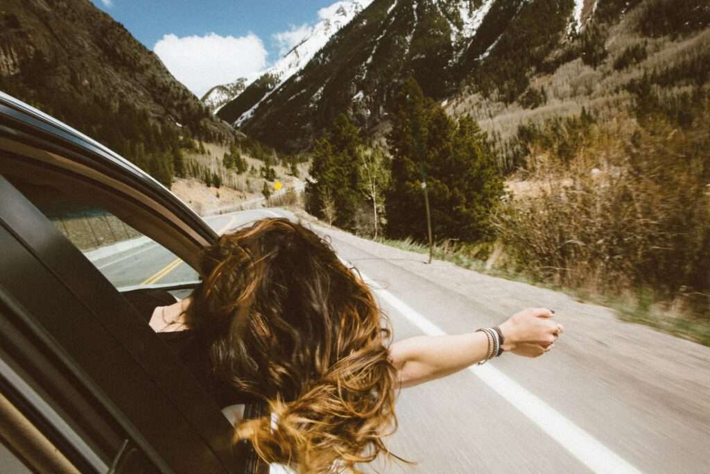A carefree girl embraces the freedom of the open road, hand out of the carwindow, surrounded by breathtaking mountain views.